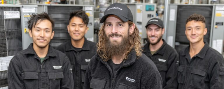 Five smiling men in matching work uniforms stand in front of industrial equipment, posing for a group photo.