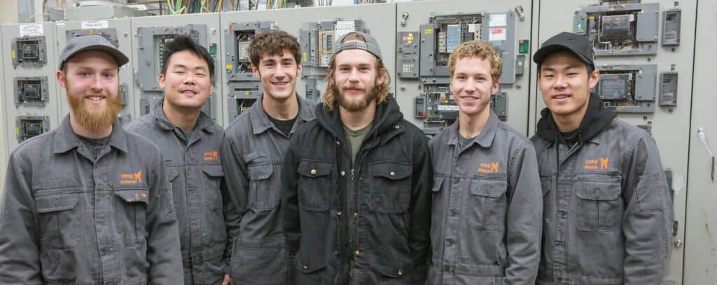 Six men in work uniforms stand smiling in front of industrial electrical panels.