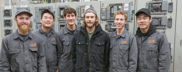 Six men in work uniforms stand smiling in front of industrial electrical panels.